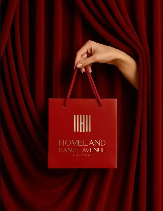Hand holding the red and gold Homeland Ranjit Avenue Amritsar retail bag against a velvet curtain